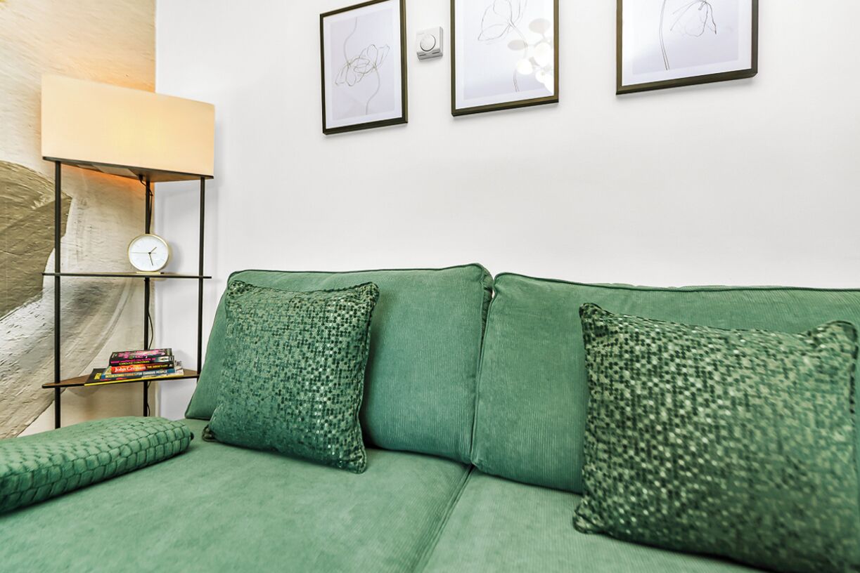 Un canapé vert agrémenté de coussins à motifs, une petite étagère avec des livres et trois œuvres d’art encadrées accrochées au mur.