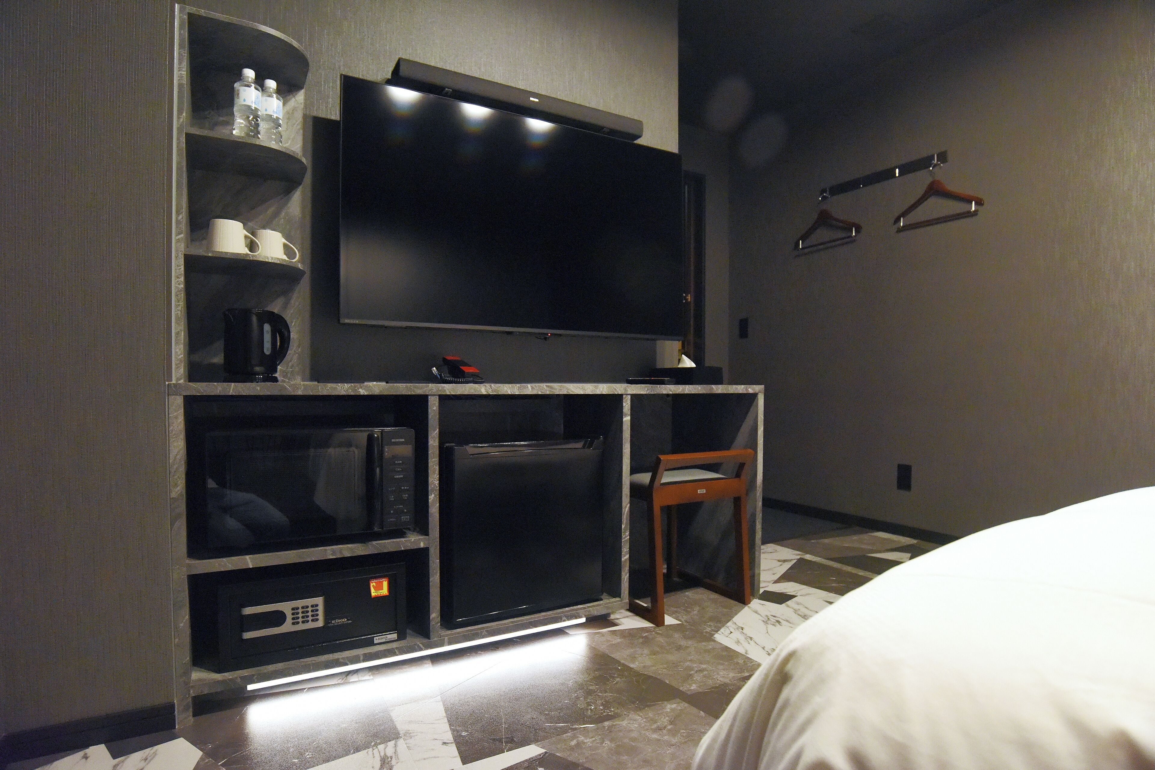 Ein Hotelzimmer mit Flachbildfernseher, Mikrowelle, Kaffeemaschine und kleinem Kühlschrank. An der Wand befinden sich zwei hölzerne Kleiderhaken und ein Sessel.