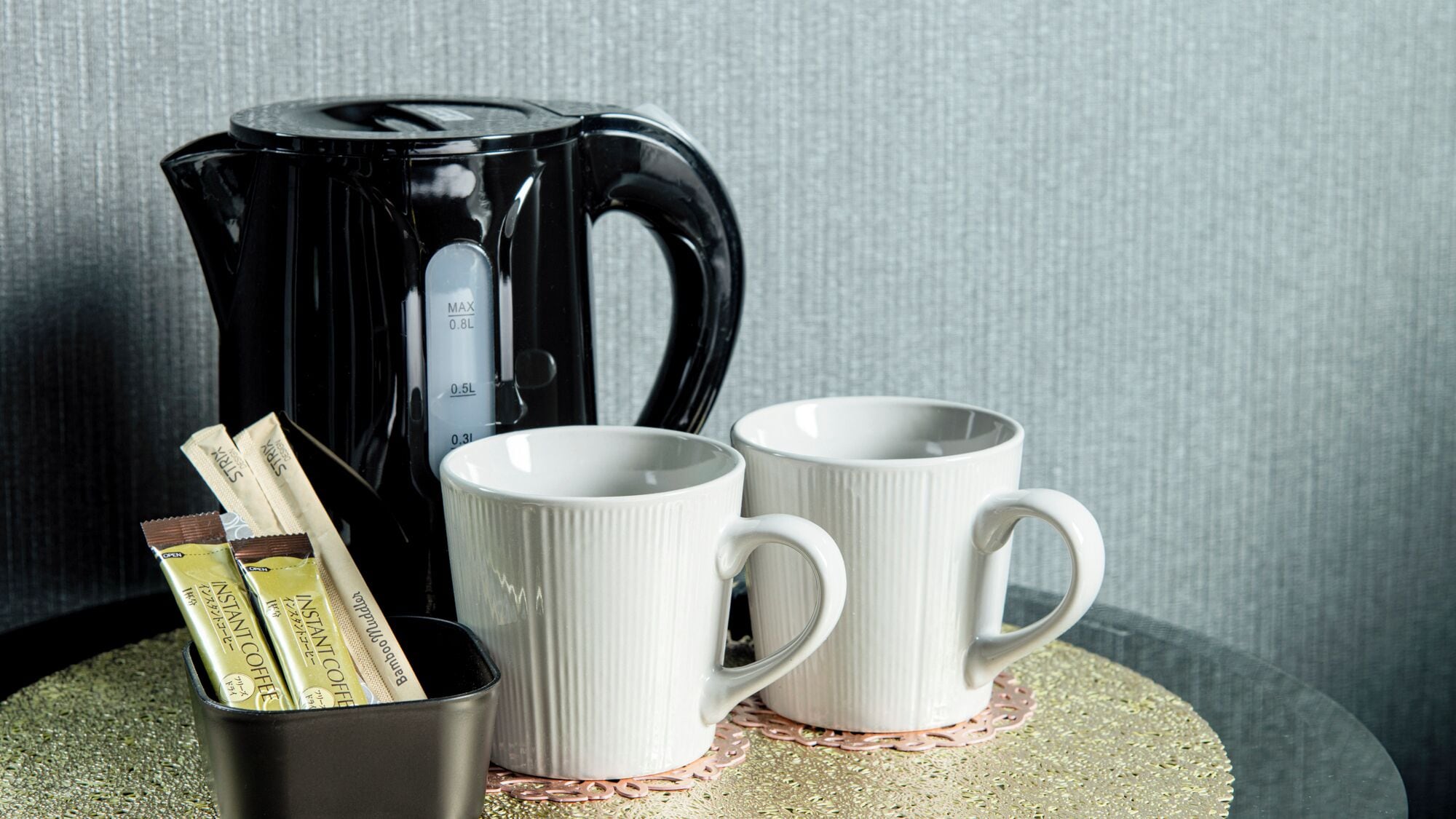 A black coffee maker, two white mugs, and instant coffee packets on a table.