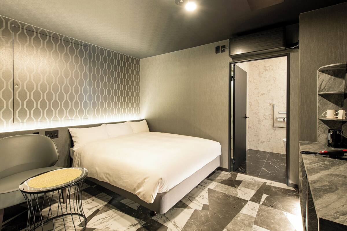 Une chambre d’hôtel moderne avec un grand lit, une petite table ronde et une chaise. On peut apercevoir la salle de bain à travers une porte ouverte.