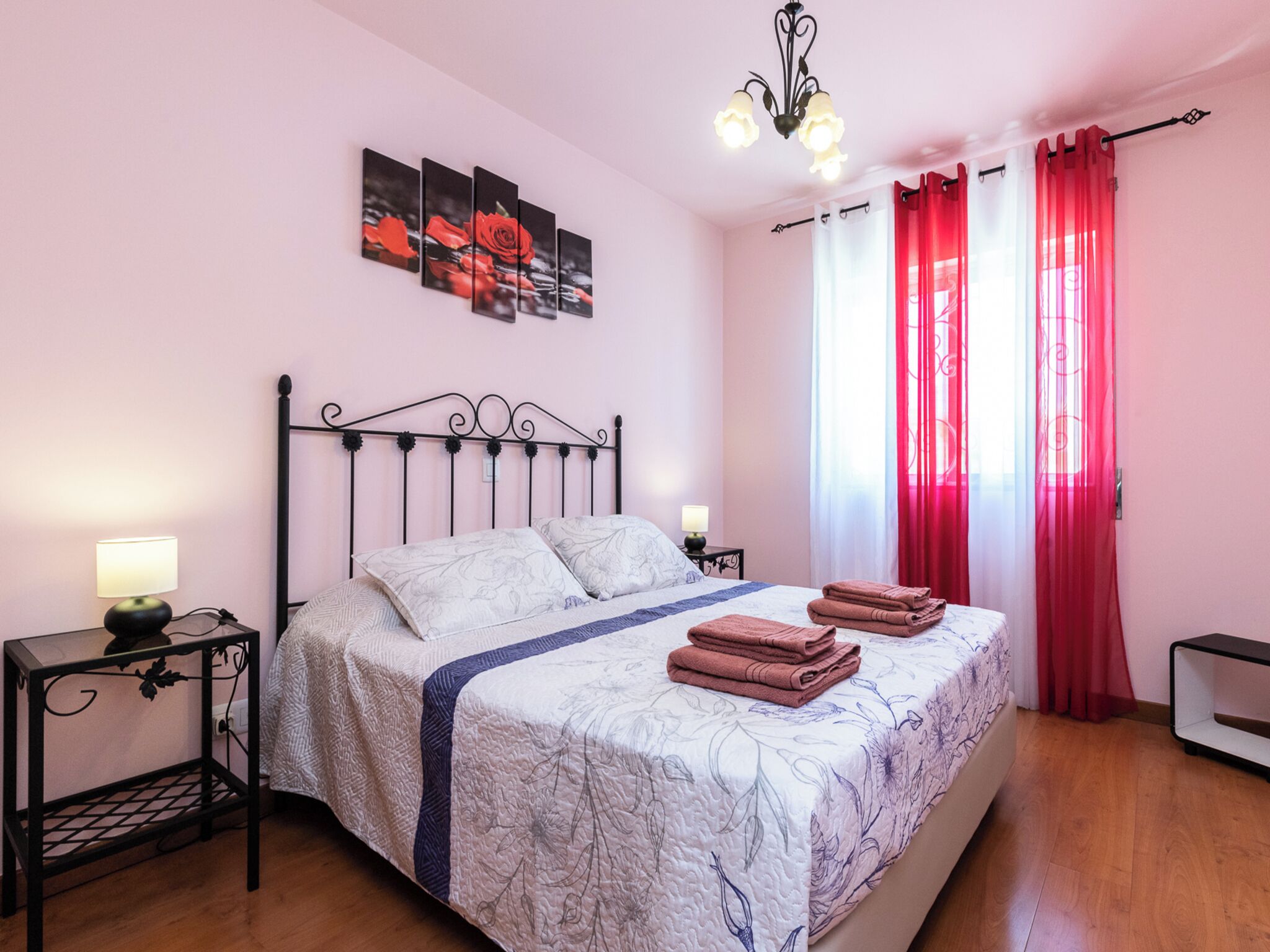 A bedroom with a bed, two bedside tables, a chandelier, and a window with red curtains.