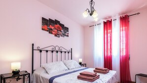 Ein Schlafzimmer mit einem Bett, zwei Nachttischen, einer Chandelieber und einem Fenster mit roten Vorhängen.