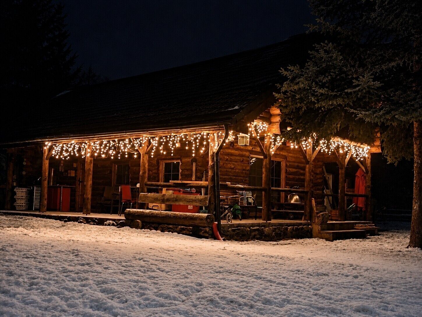 Una cabaña de troncos con un porche decorado con luces colgantes, suelo cubierto de nieve y una valla de madera.