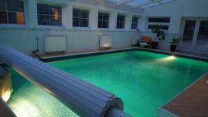 An indoor swimming pool with a wooden deck, a covered patio area, and a view of the surrounding building.