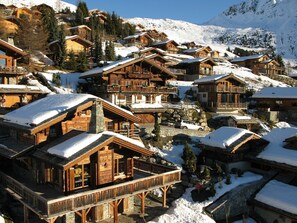 Een groepje houten chalets met balkons, voor een besneeuwd berglandschap.
