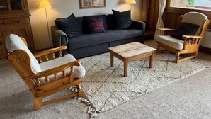 A cozy living room with a dark grey sofa, two wooden armchairs, a small wooden coffee table, and a large window offering a mountain view. - MAGNIFICENT VIEWS FROM THE HEART OF VERBIER-NEXT TO THE NEW SPORTS CENTER (Val de Bagnes)