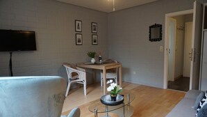 A modern living room with a flat-screen TV, a dining table with chairs, and a glass coffee table with a flower vase.