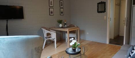 A modern living room with a flat-screen TV, a dining table with chairs, and a glass coffee table with a flower vase.