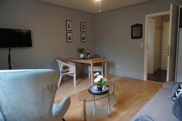 Uma sala de estar moderna com uma TV de tela plana, uma mesa de jantar com cadeiras e uma mesa de centro de vidro com um vaso de flores.