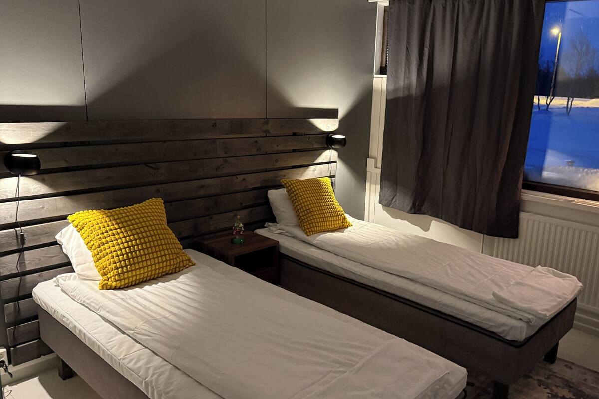 A room with two single beds, wooden headboards, a nightstand with a lamp, a window with a view of a snowy landscape, and a radiator.