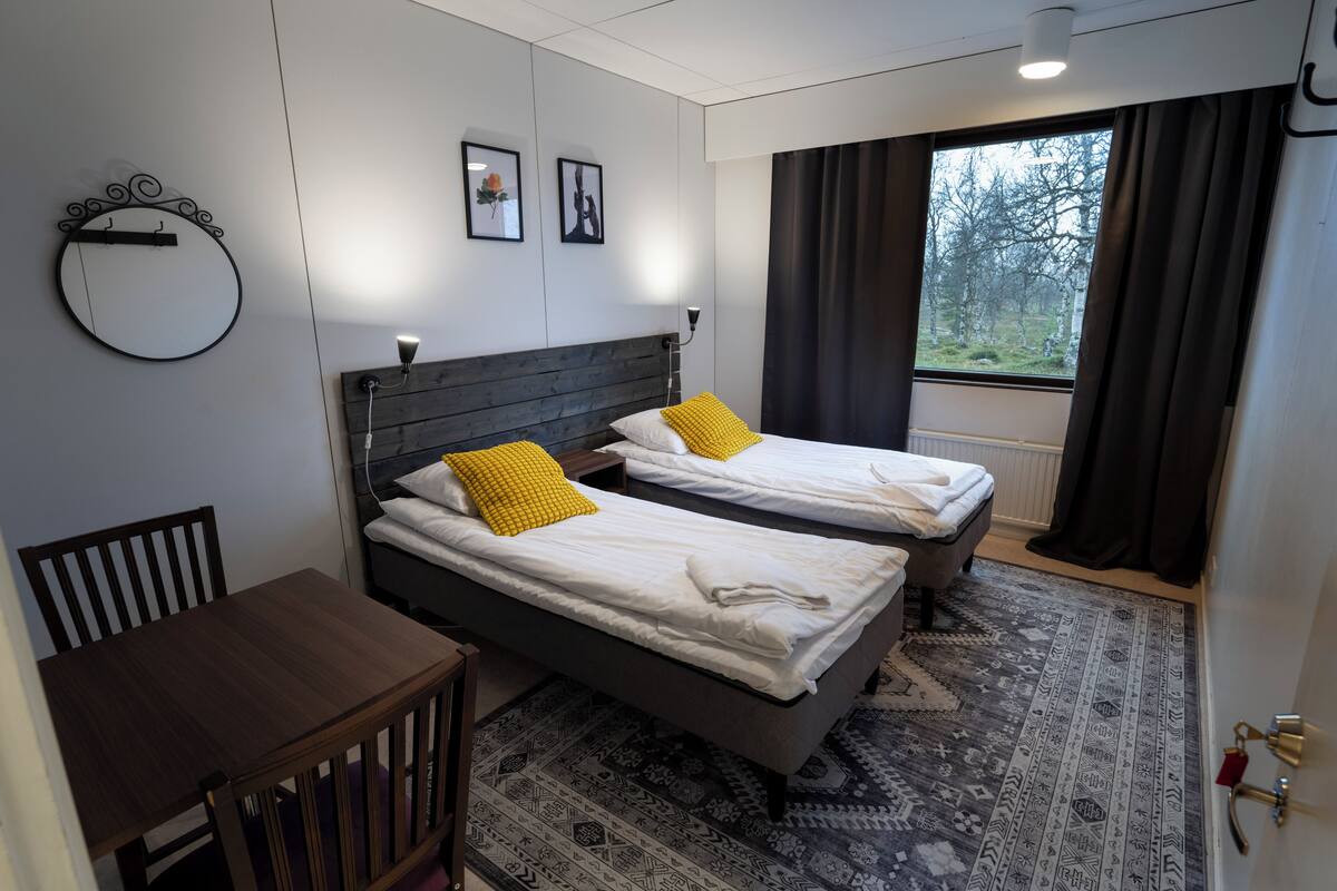 A hotel room with two beds, a wooden table, a round mirror, and a view of trees through the window.