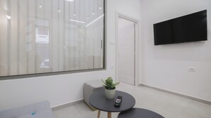 Quarto moderno com uma janela ampla, televisão de tela plana na parede, uma mesinha redonda com planta em vaso e controle remoto, e um sofá cinza.