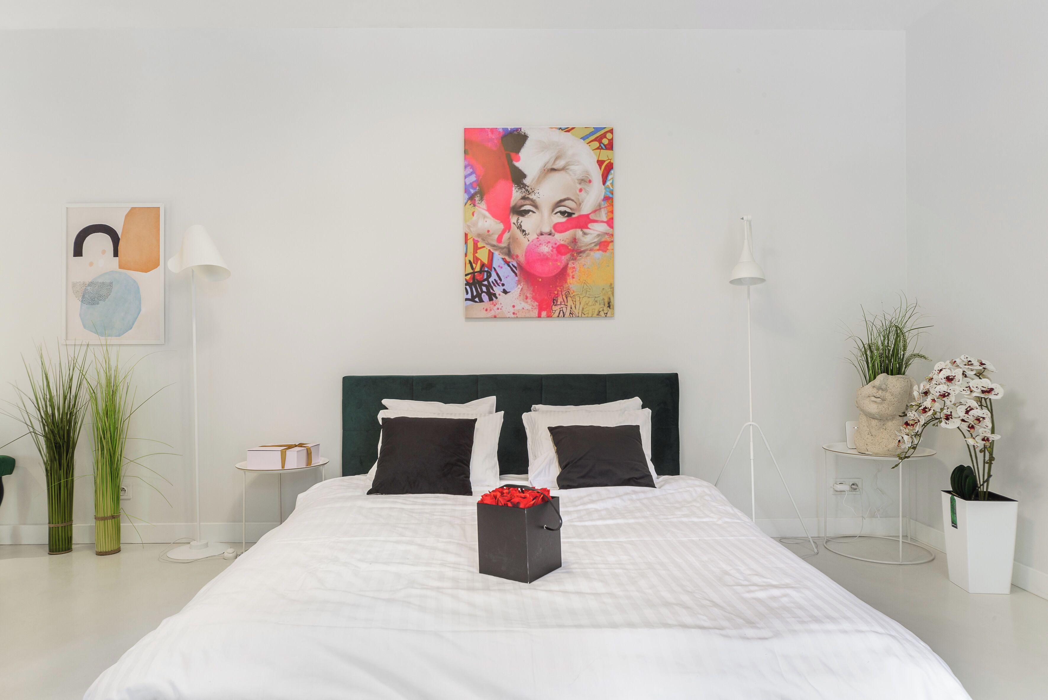 A modern bedroom with a large bed, two black pillows, a white bedside table, a small white table, a green plant, and a potted orchid.