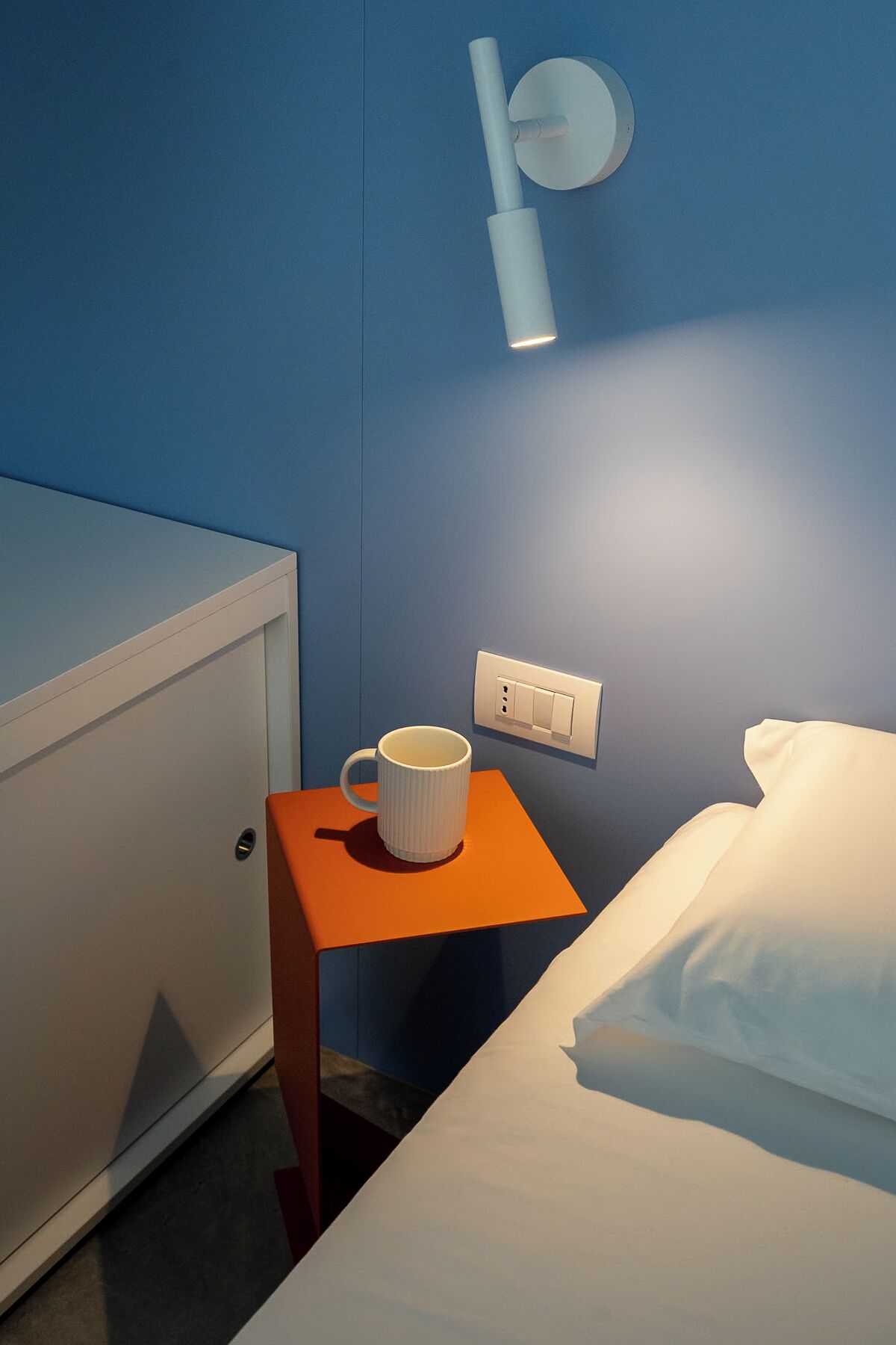 Une chambre d’hôtel avec un lit, une table de chevet sur laquelle se trouve une tasse blanche, une lampe fixée au mur et une prise de courant.