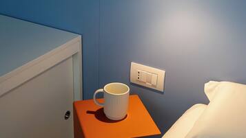 Une chambre d’hôtel avec un lit, une table de chevet sur laquelle se trouve une tasse blanche, une lampe fixée au mur et une prise de courant.