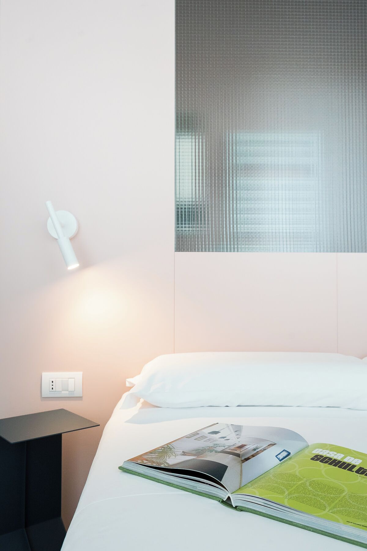 Une chambre d’hôtel avec un lit, une table de chevet, une lampe fixée au mur et une fenêtre avec des stores.