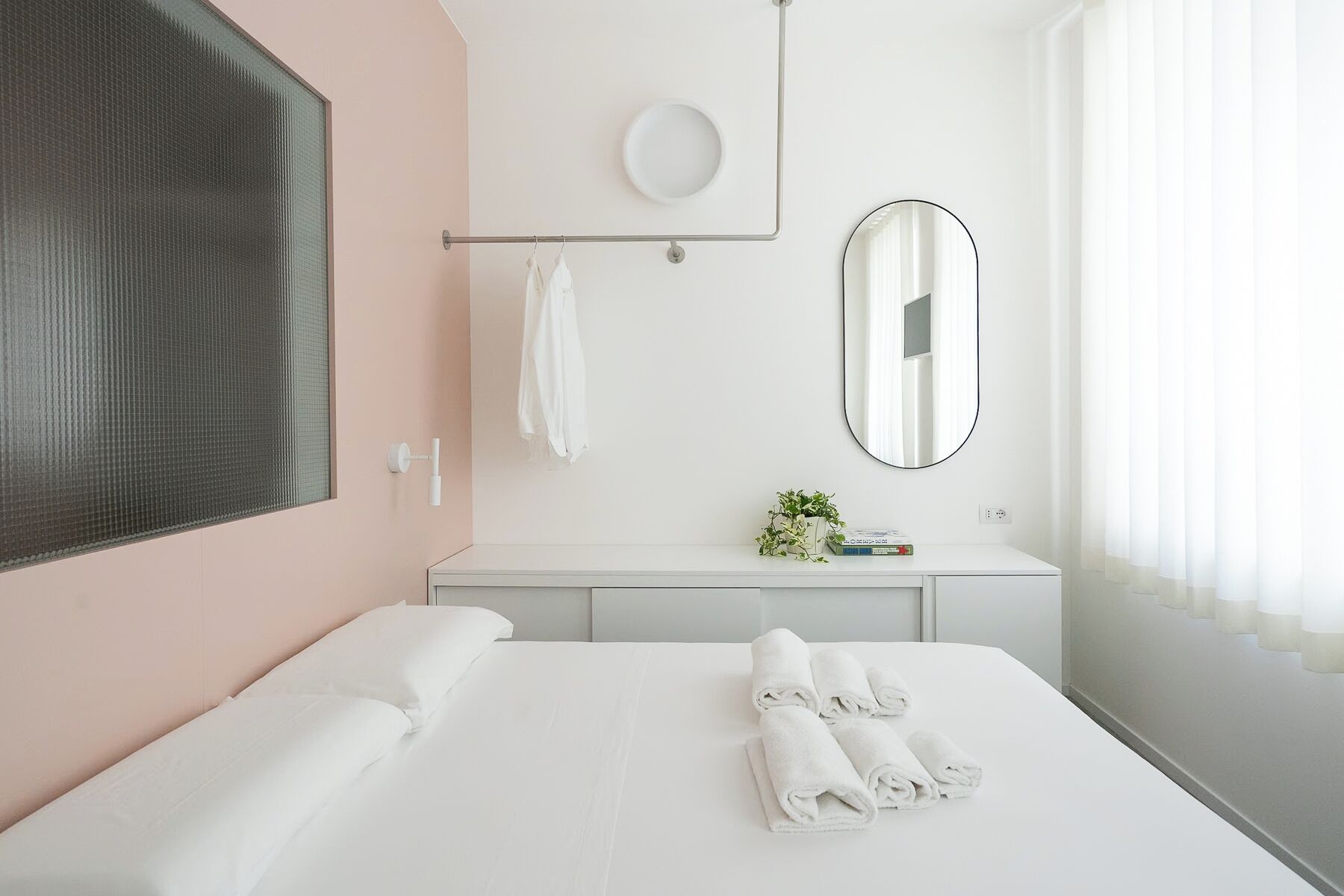 Una camera d'albergo con un letto grande, un comodino con uno specchio, un appendi asciugamani e una finestra con tende.