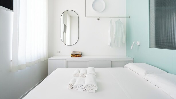A modern hotel room with a large bed, a bedside table with a mirror, a small shelf with toiletries, and a window with blinds.