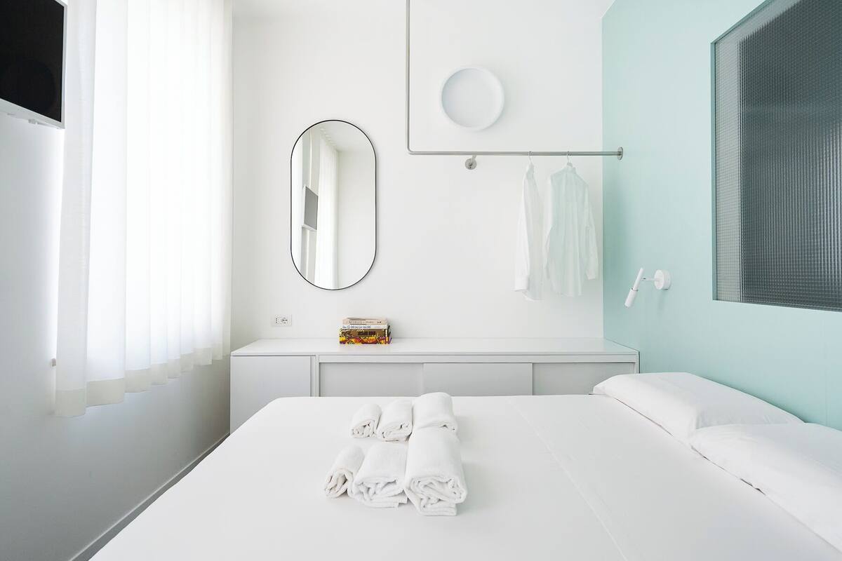 A modern hotel room with a large bed, a bedside table with a mirror, a small shelf with toiletries, and a window with blinds.