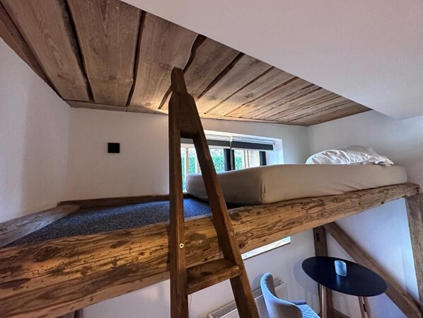 Un lit superposé avec une échelle en bois, une petite table et une fenêtre avec des stores.