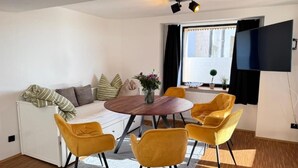 A modern living room with a sofa, a dining table with four chairs, a flat-screen TV mounted on the wall, and a window with curtains. - SunDeck Apartment (Am Ettersberg)