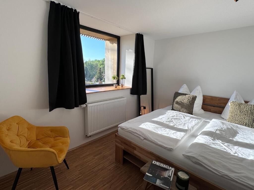 A bedroom with a bed, a yellow chair, a window with curtains, a radiator, and a magazine on a table.