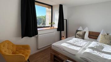 A bedroom with a bed, a yellow chair, a window with curtains, a radiator, and a magazine on a table.