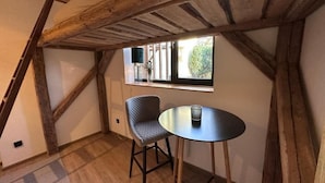 A cozy room with exposed wooden beams, a round table, a cushioned chair, and a window with a view of greenery. - SunDeck Apartment (Am Ettersberg)