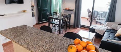 Een moderne keuken met een granieten aanrecht, een eethoek met een glazen tafel en stoelen, en een zithoek met een bank en een televisie.