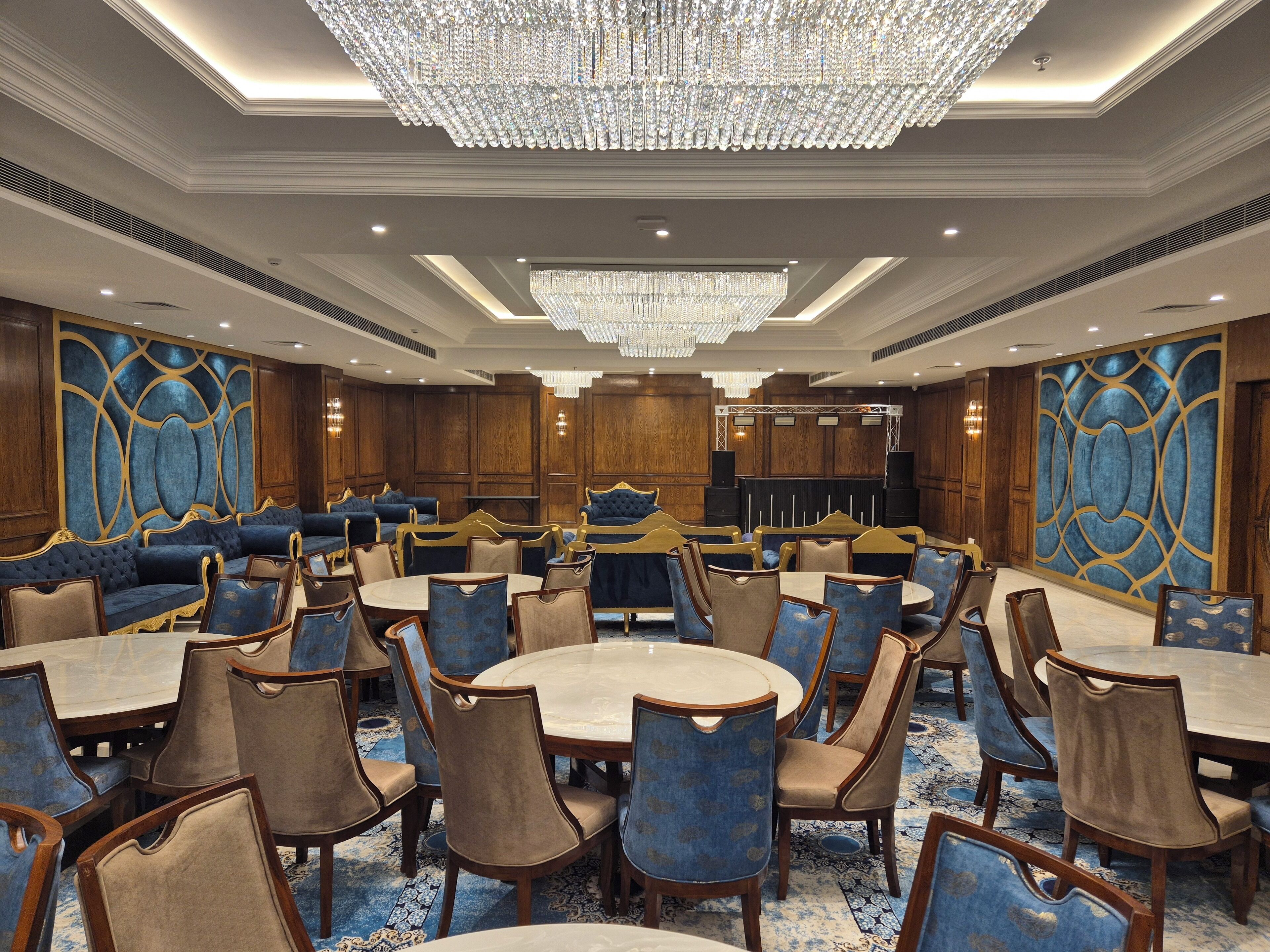 A spacious hotel lobby with round tables and blue upholstered seating, a large chandelier, and a stage with speakers.