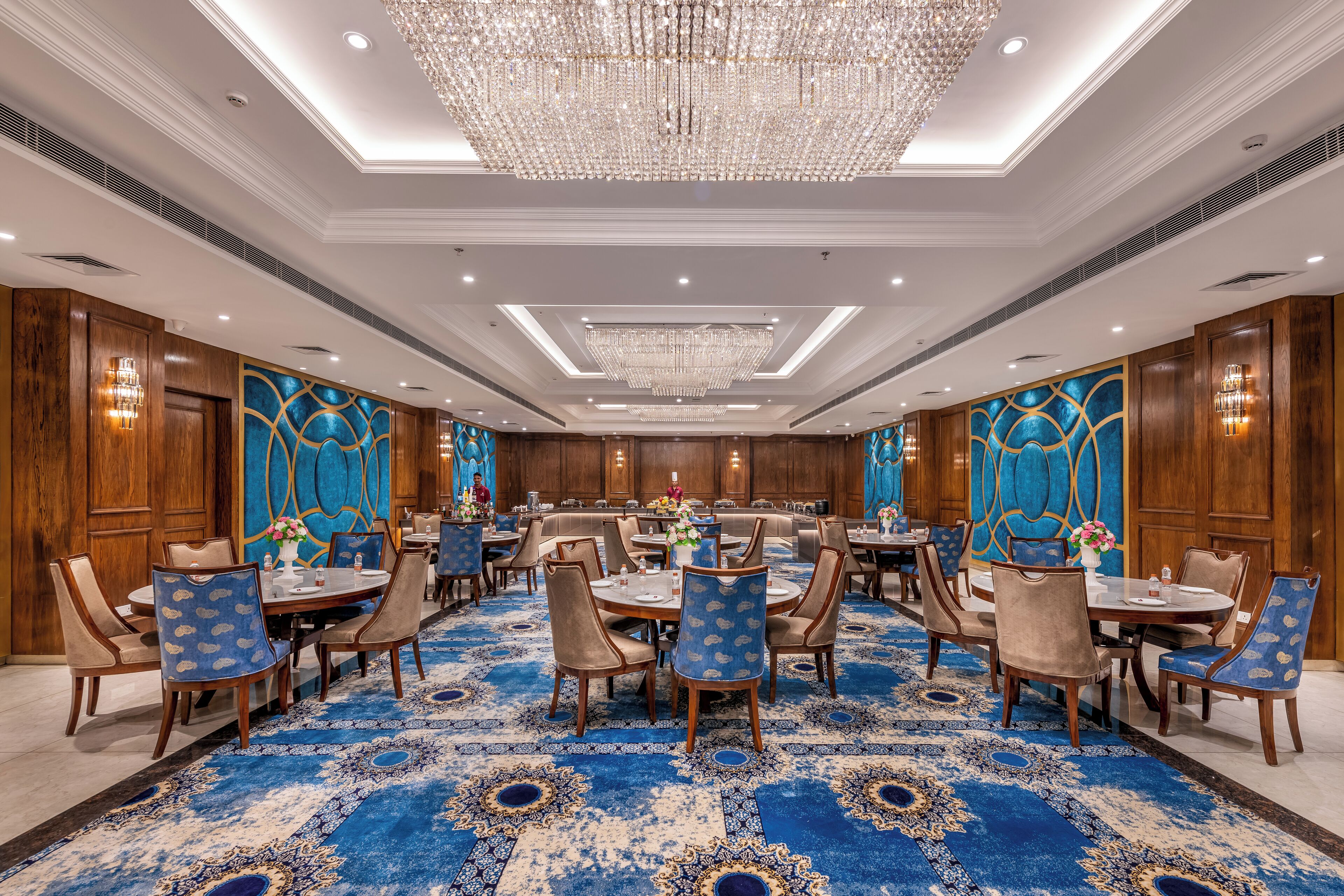 A spacious banquet hall with round tables, wooden paneling, and a large blue patterned carpet.