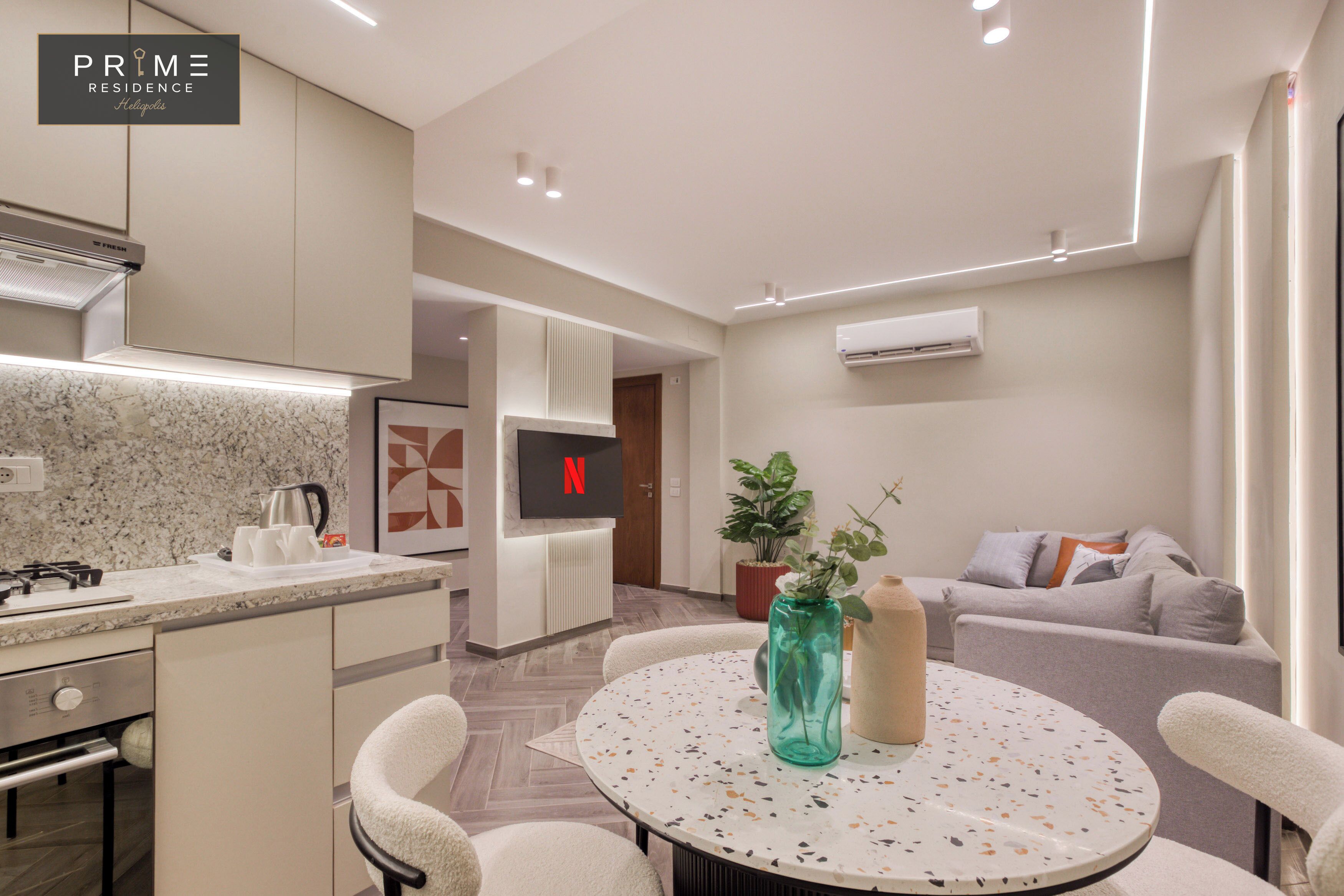 Una cocina moderna con estufa integrada, cafetera y televisor montado en la pared. En la zona de estar, hay una mesa redonda con sillas y un sofá con cojines.