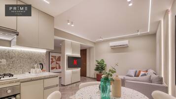 Una cocina moderna con estufa integrada, cafetera y televisor montado en la pared. En la zona de estar, hay una mesa redonda con sillas y un sofá con cojines.