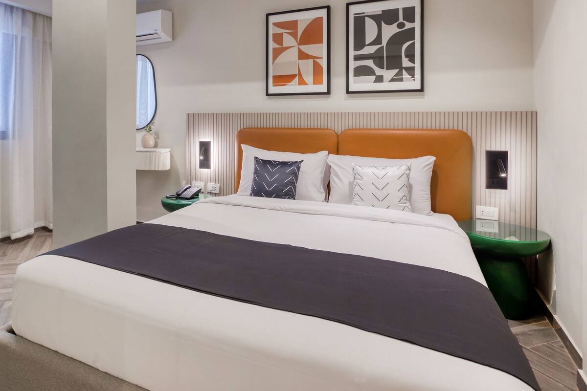 Una habitación de hotel con una cama grande, una mesita de noche con lámpara, un espejo y arte abstracto en la pared.