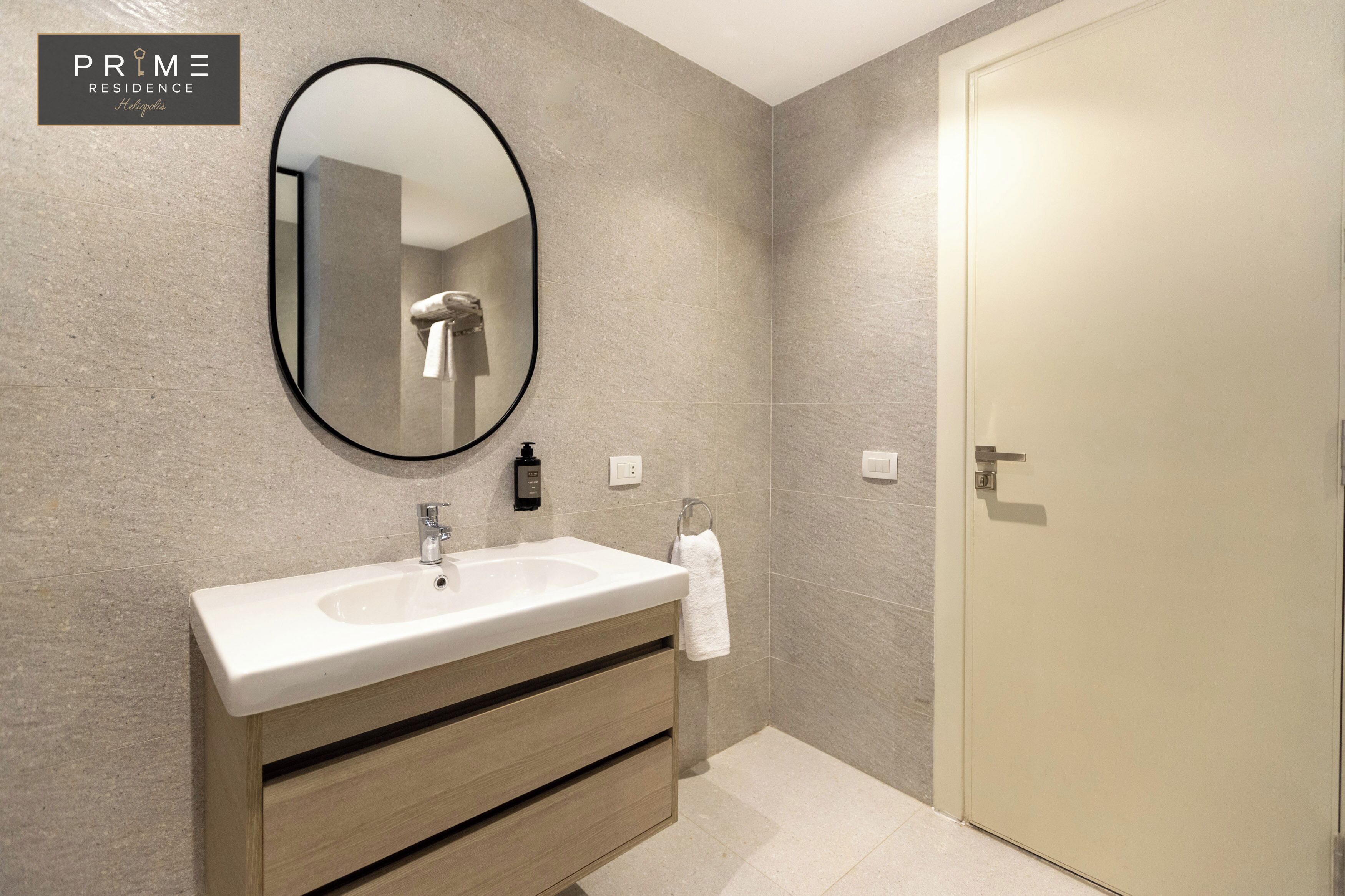 A modern bathroom with a large oval mirror, a white sink, wooden vanity, and a beige door.