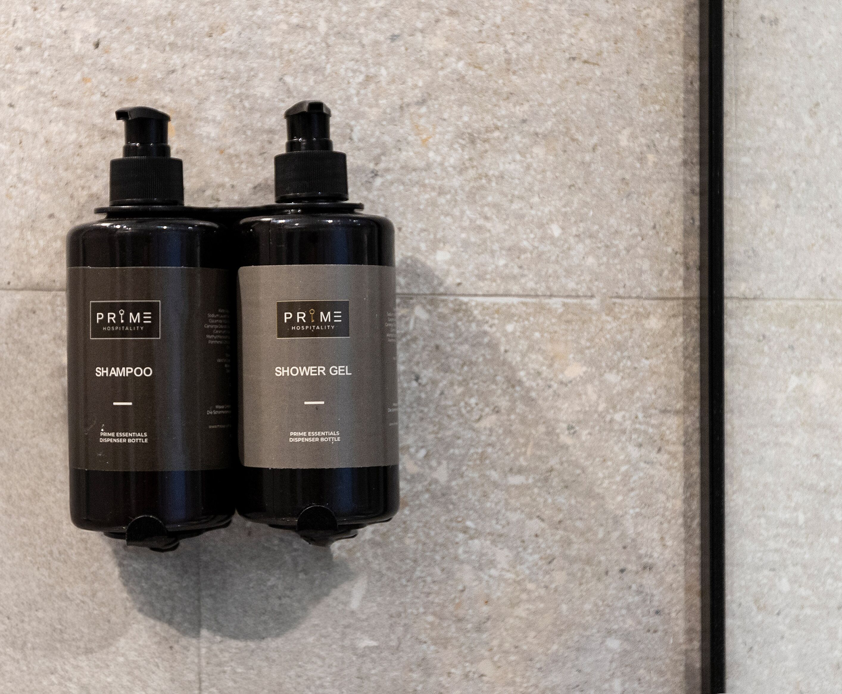 Two black Prime Hospitality shampoo and shower gel bottles mounted on a textured wall.