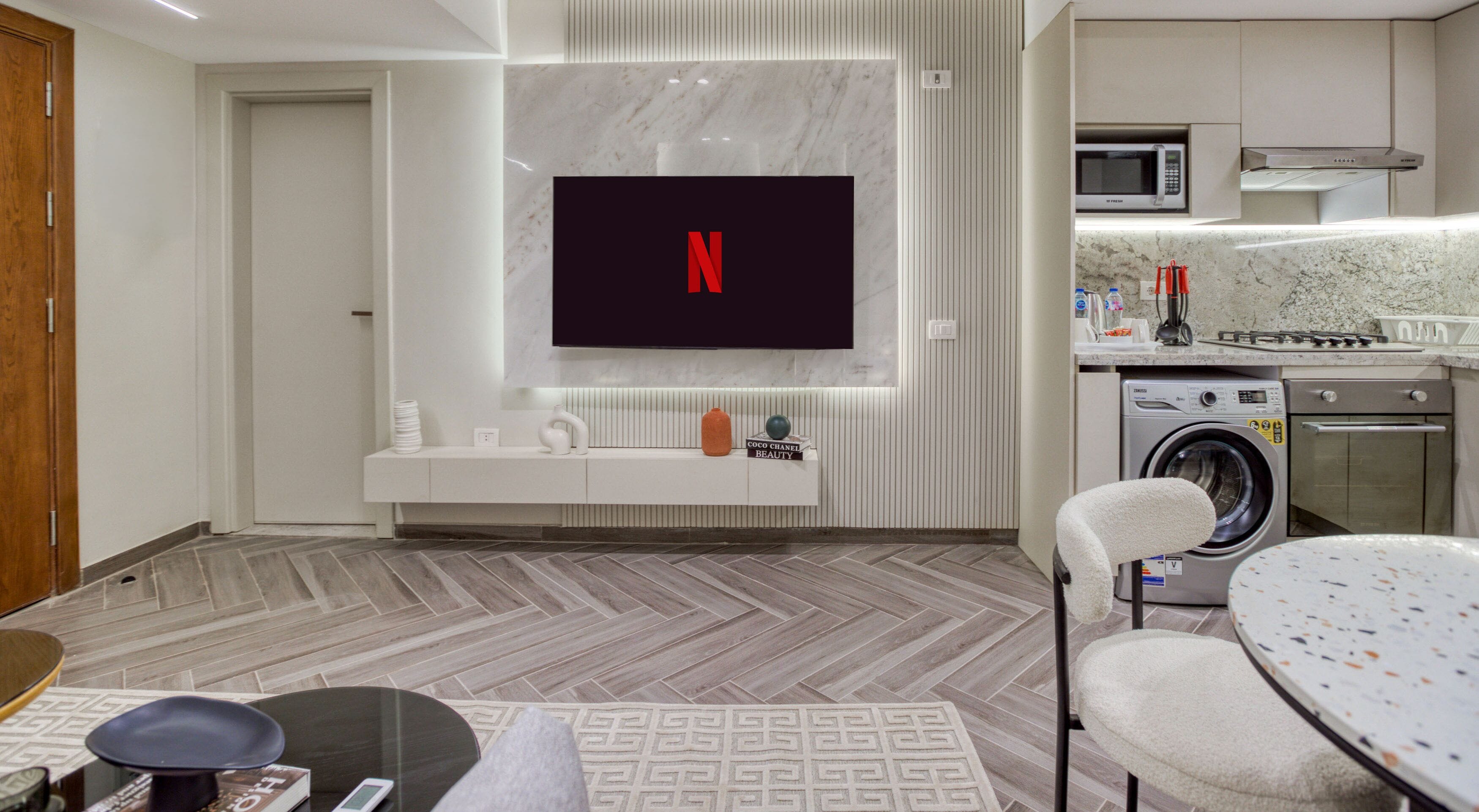 Een moderne woonkamer met een flatscreen televisie aan de muur, een ingebouwde wasmachine en droger, een keukenhoek met een magnetron, en een visgraatvloer.
