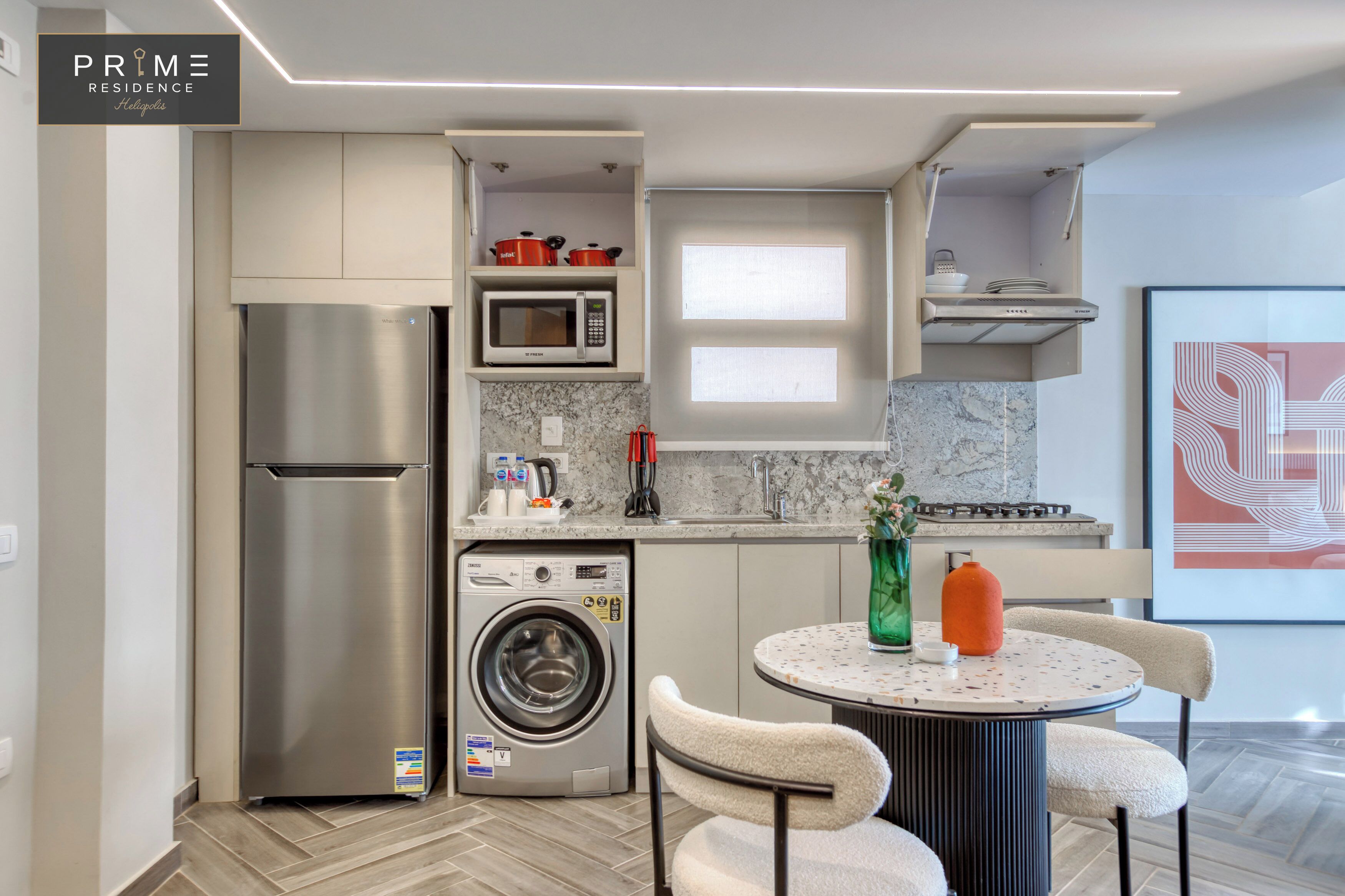 A compact kitchen with a stainless steel refrigerator, a microwave, and a built-in washing machine. There is a dining area with a round table and chairs.