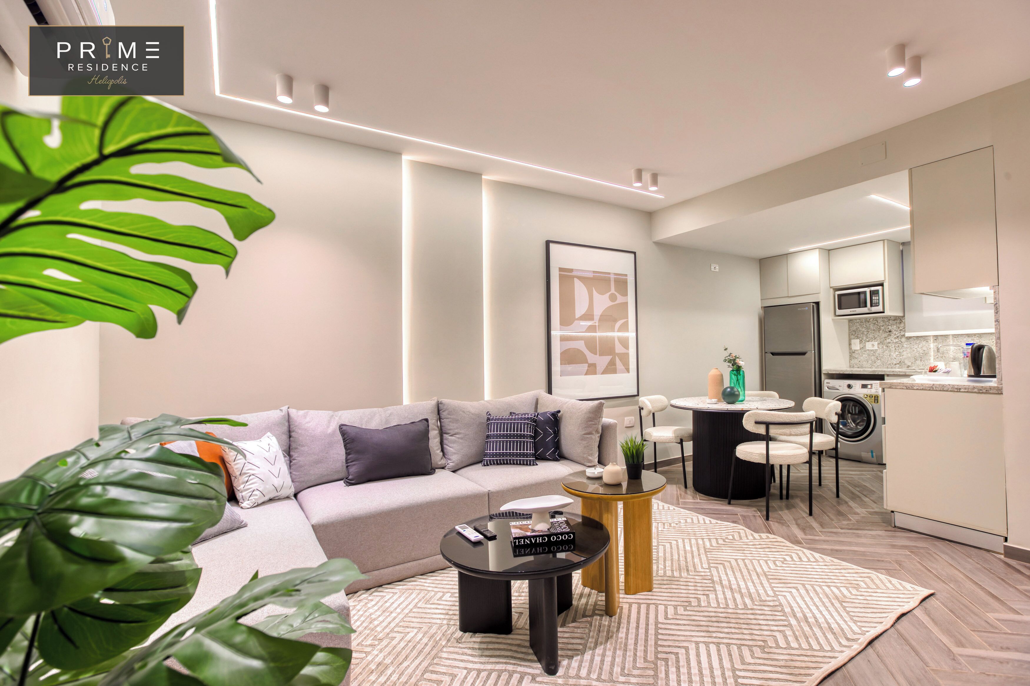A modern living room with a sofa, coffee table, and a dining area with chairs. There is a kitchen in the background with appliances and a microwave.