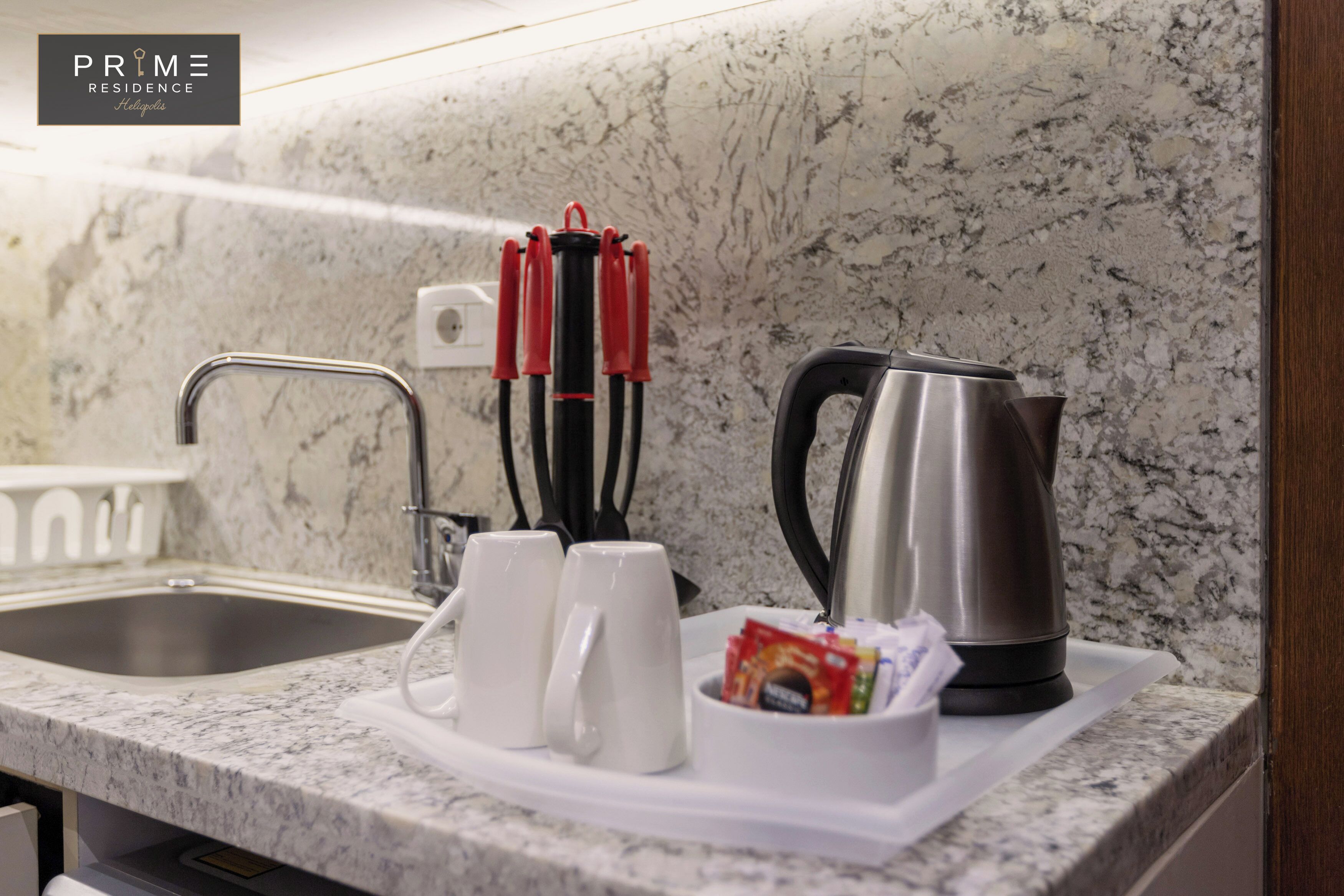 Una encimera de cocina con un lavabo, dos tazas blancas, una tetera y utensilios de cocina con mangos rojos.