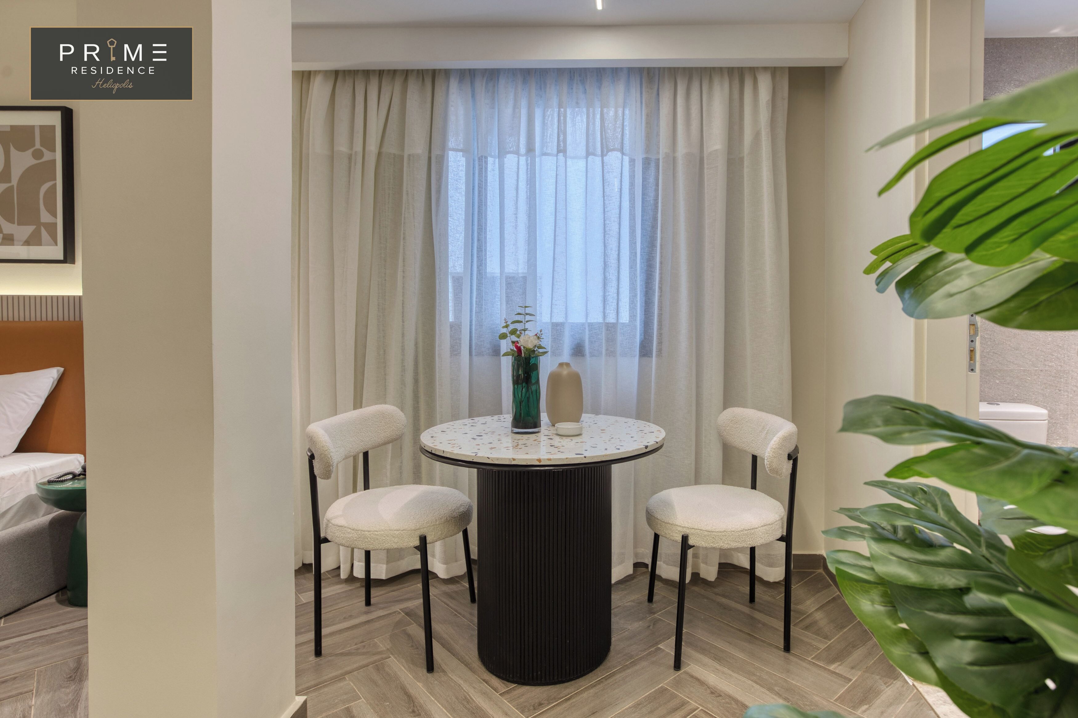 A modern room with a round table, two chairs, a vase with flowers, and a large window with sheer curtains.