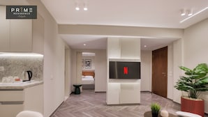 Moderni hotellihuone, jossa on pieni keittiö, taulutelevisio, sänky ja ruukkukasvi.