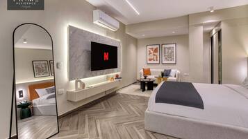 Quarto de hotel moderno com uma cama grande, uma televisão montada na parede, um espelho e uma área de estar com uma mesinha.