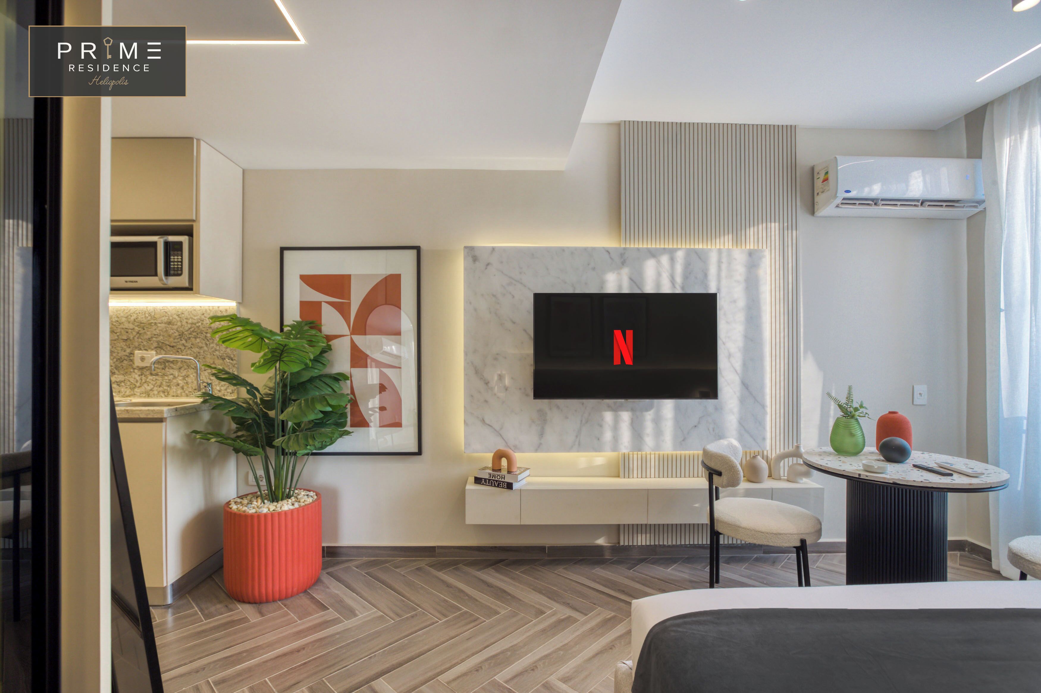 Una habitación de hotel moderna con televisor de pantalla plana en la pared, cocina integrada, zona de estar con mesa redonda y una planta en maceta.