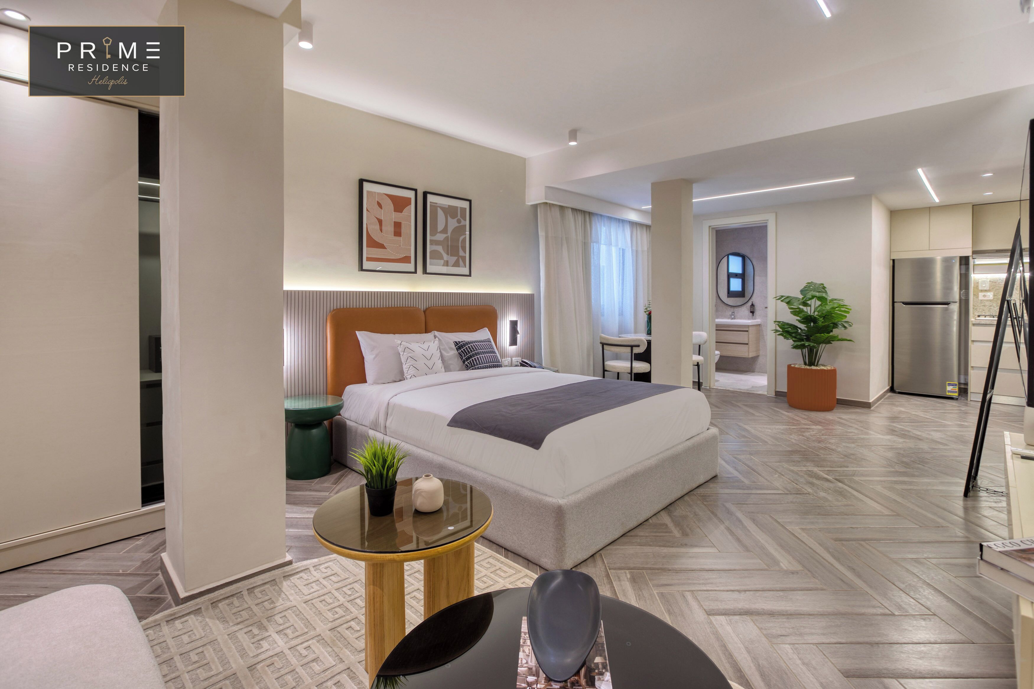 Une chambre d’hôtel moderne avec un grand lit, un petit bureau, une chaise, une plante en pot et une kitchenette.
