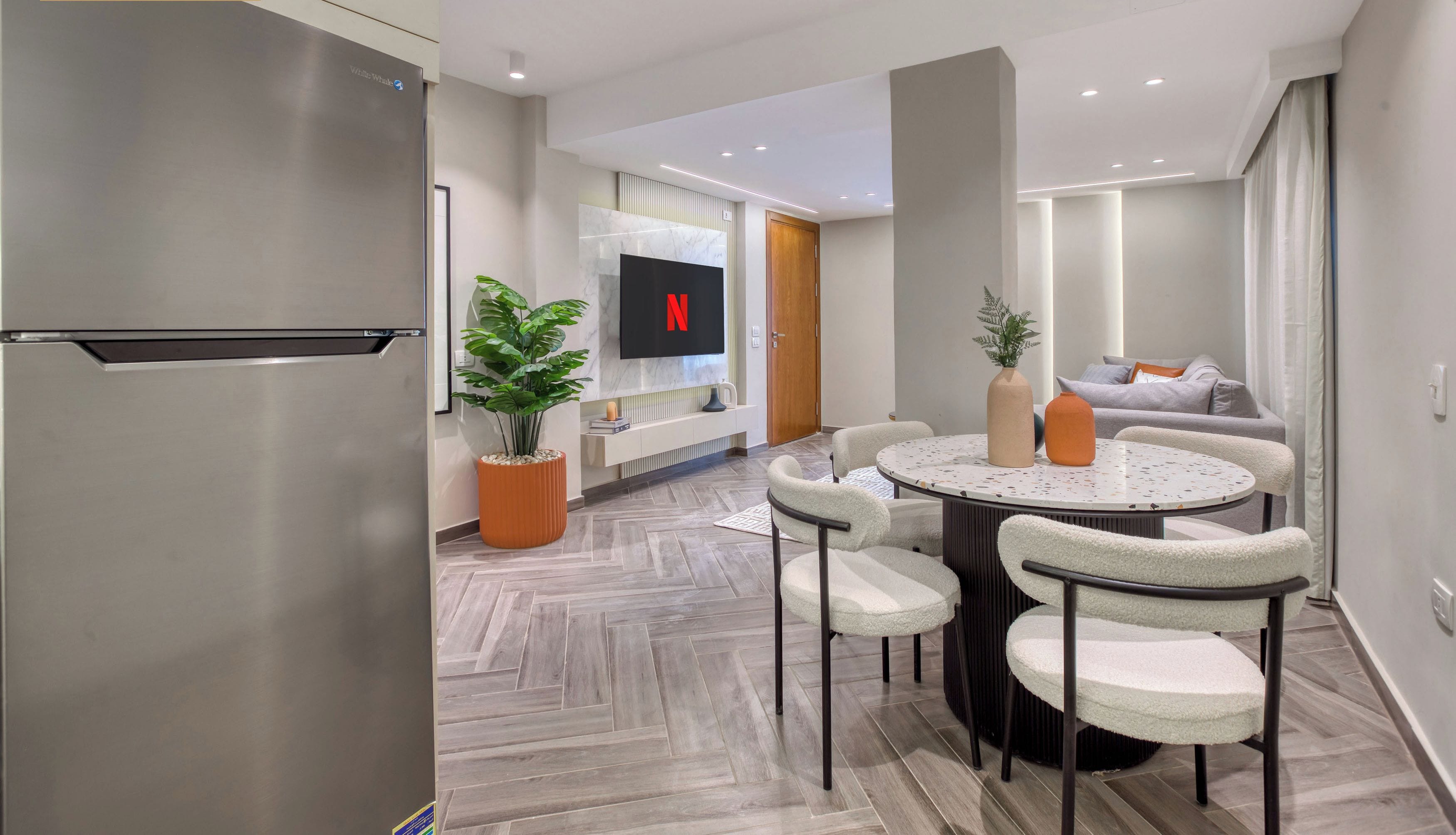 A modern living room with a TV, a dining area with a round table and chairs, and a kitchen with a refrigerator.