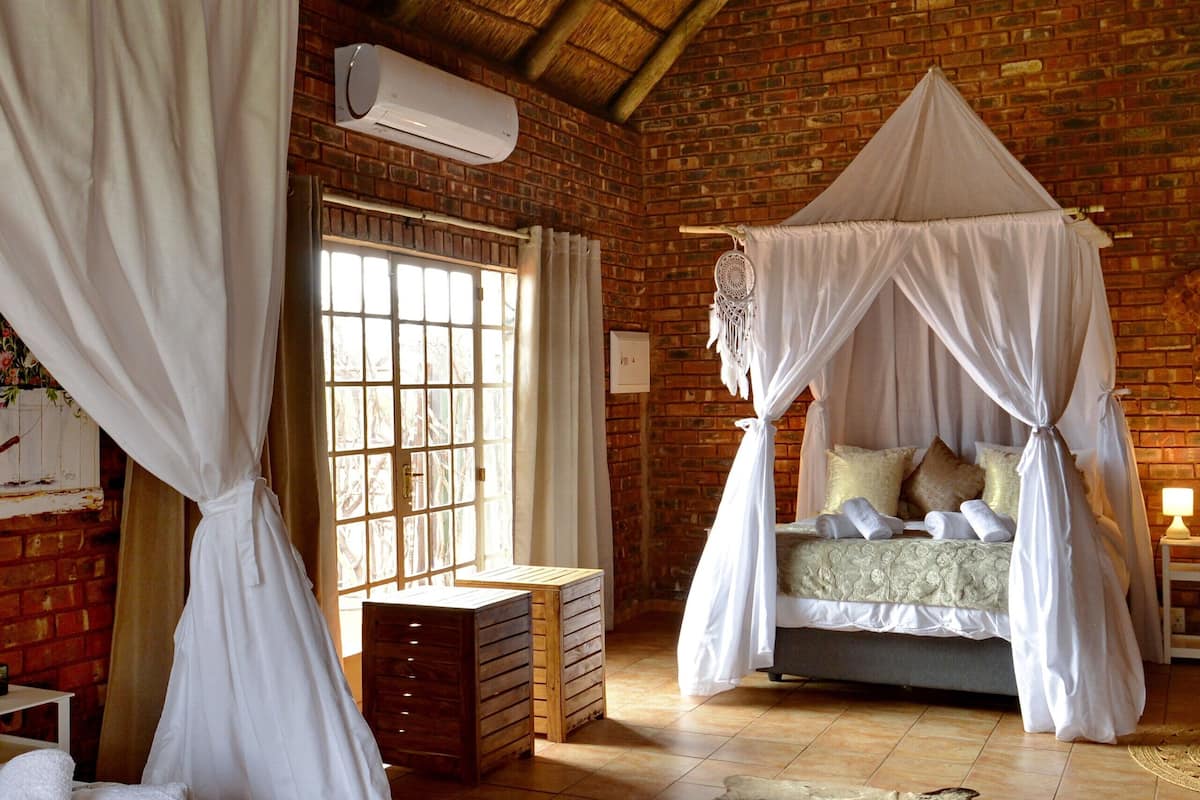 Una habitación con pared de ladrillo, techo de paja, cama con dosel, ventana con cortinas y alfombra en el suelo.