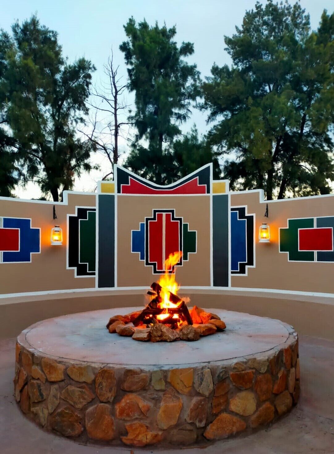 A stone fire pit with a fire burning in the center, surrounded by a decorative wall with geometric patterns and lights.