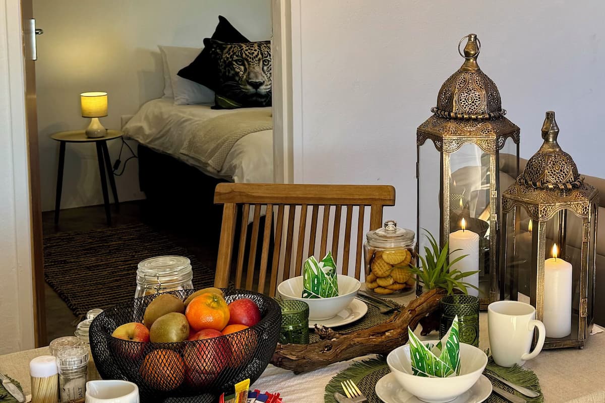 Una mesa de comedor dispuesta con frutas, sobres de azúcar y velas, con una cama y una lámpara al fondo.