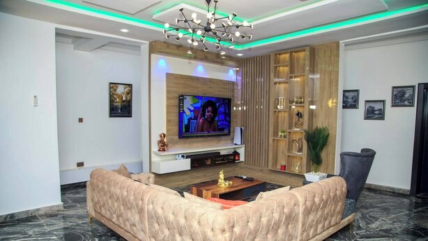 A modern living room with a large sectional sofa, a flat-screen TV mounted on a wooden wall, and a decorative ceiling light. - The Captains Apartment Abuja (Abuja)